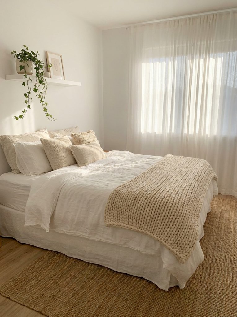 White aesthetic bedroom with layered white linen bedding neutral throw pillows chunky knit blanket sheer curtains trailing plant on floating shelf calm effortlessly beautiful